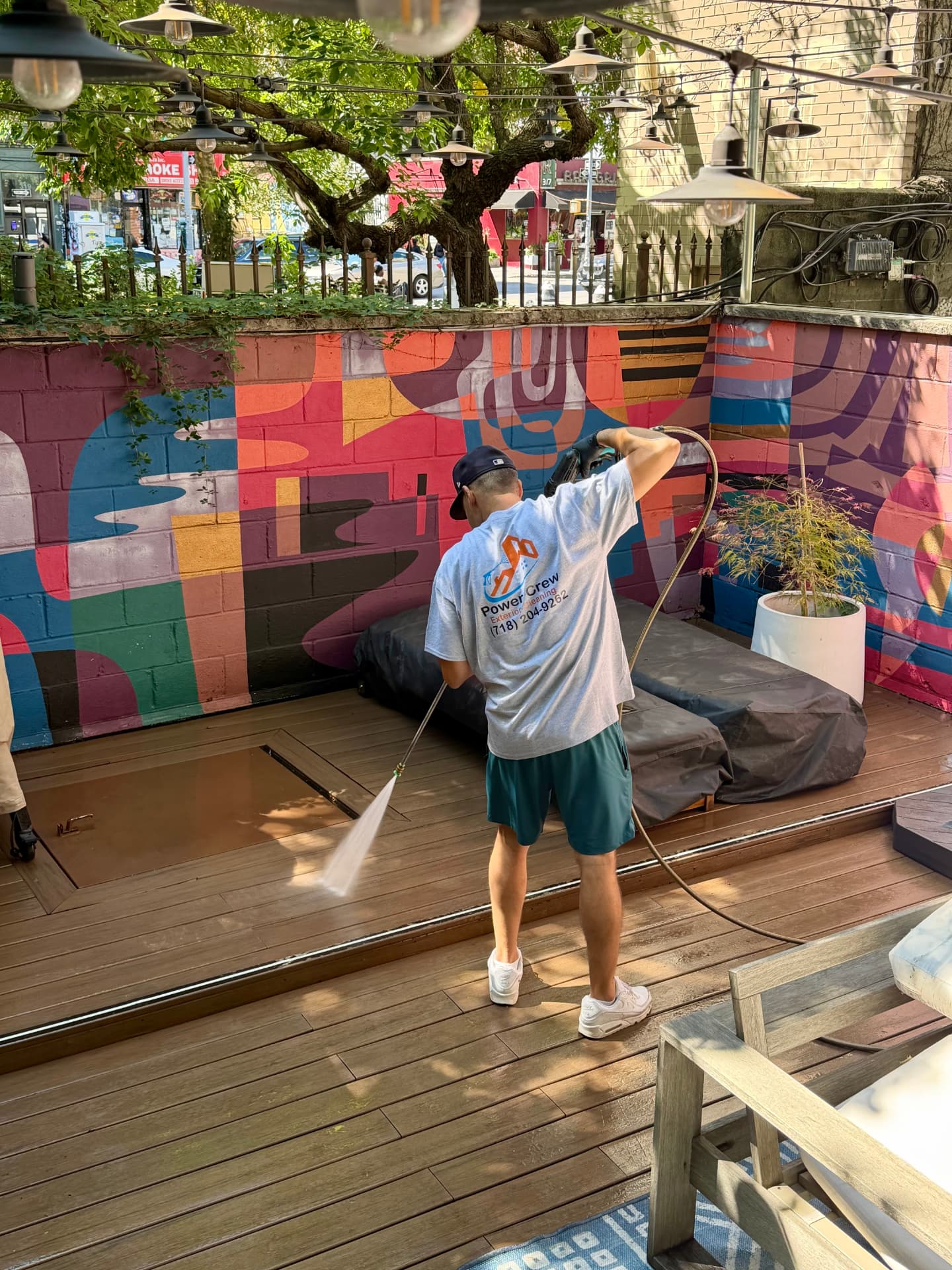 Power Crew technician pressure washing a backyard patio deck in front of a colorful mural in NYC